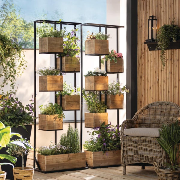 Modernes Pflanzregal aus schwarzem Metall mit versetzt angeordneten Holzkisten auf einer hellen Holzterrasse mit Bambuswand und Korbstuhl.