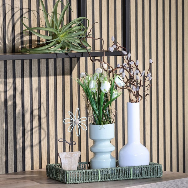 Dekorative Szene mit Pflanzen, Vasen und einem Metallornament auf einem geflochtenen Tablett vor einer Holzwand