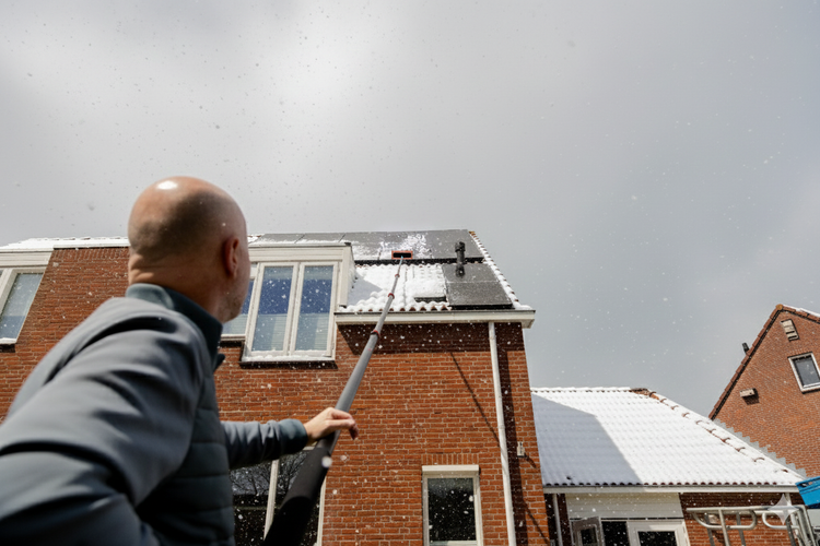 Eine Person reinigt mit einem Teleskopstiel und einer Bürste Schnee von Solarmodulen auf einem Dach.