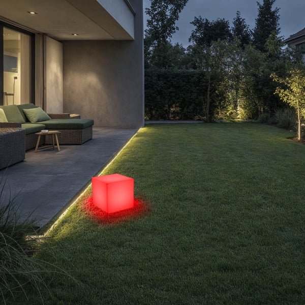 Beleuchteter LED-Würfel im Garten auf einer Rasenfläche neben einer Terrasse mit modernen Loungemöbeln bei Dämmerung.