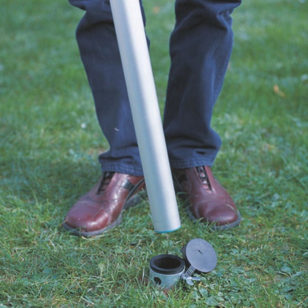 Eine Person führt ein Metallrohr in eine im Rasen installierte Bodenhülse mit geöffnetem Deckel ein.