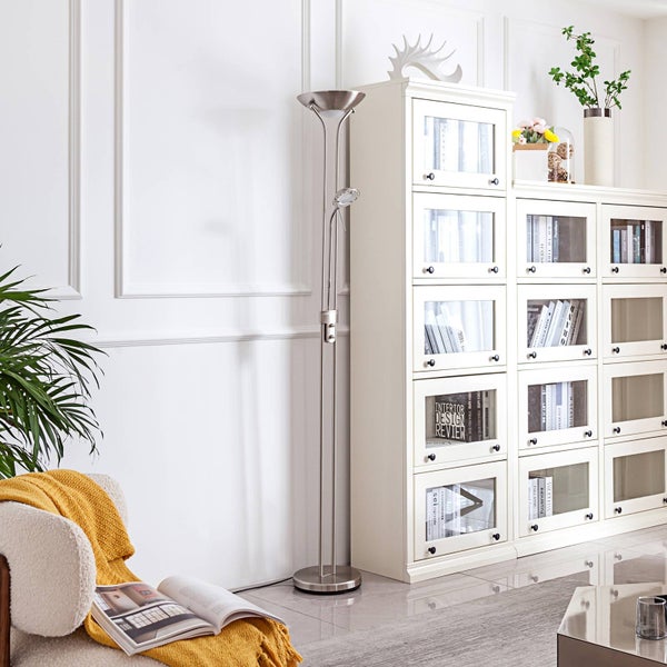 Moderne Stehleuchte aus Metall mit Deckenfluter und Lesearm in einem hellen Wohnzimmer neben einem weißen Bücherregal und einem Sessel.
