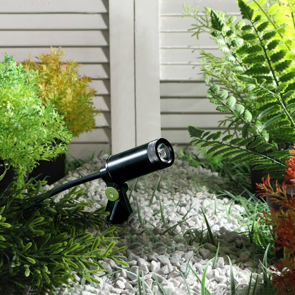 Schwarzer Gartenstrahler platziert auf weißen Kieselsteinen inmitten von grünen Pflanzen und Farnen vor einer hellen Holzwand mit Lamellen.