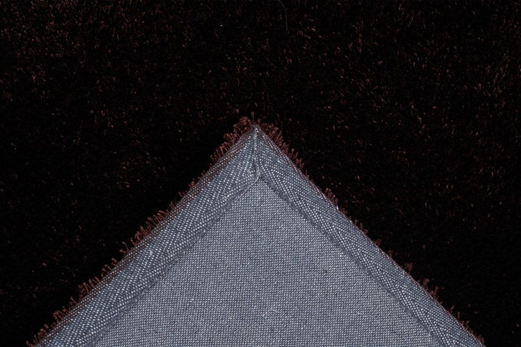 Dunkelbrauner Hochflor-Teppich mit Detailansicht der gewebten Rückseite an einer umgeklappten Ecke.
