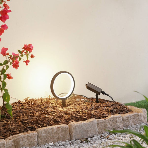 Moderner kreisförmiger LED-Gartenstrahler und Transformator in einem Beet mit Rindenmulch, Steineinfassung und rosa Blüten.