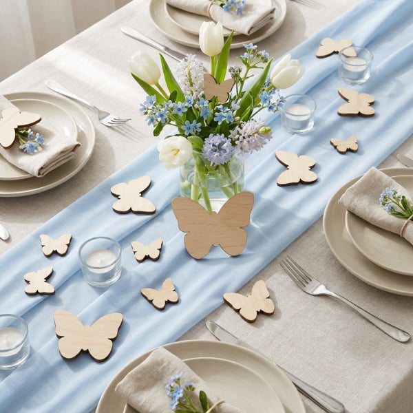 Festlich gedeckter Tisch mit Frühlingsdekoration aus Holzschmetterlingen, Blumenstrauß in Glasvase, Kerzengläsern und Leinentischdecke.
