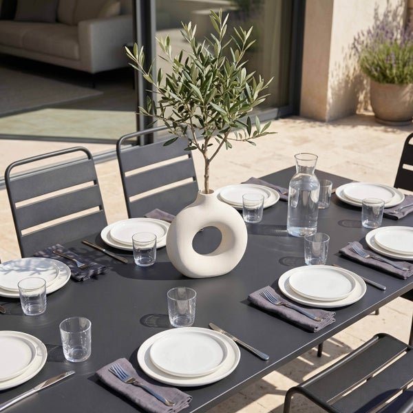 Gedeckter Gartentisch aus Metall auf einer hellen Terrasse mit modernen Stühlen, weißem Geschirr und einer dekorativen Vase mit Olivenzweig.