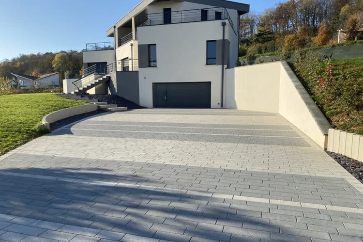 Moderne Hofeinfahrt mit rechteckigen Pflastersteinen aus Beton in gestreiftem Muster vor einem Wohnhaus mit Garage.