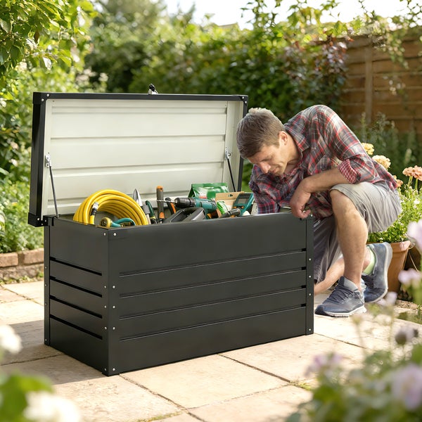 Ein Mann blickt in eine geöffnete schwarze Gartenbox aus Metall auf einer Terrasse, gefüllt mit Gartenschläuchen und Werkzeugen.