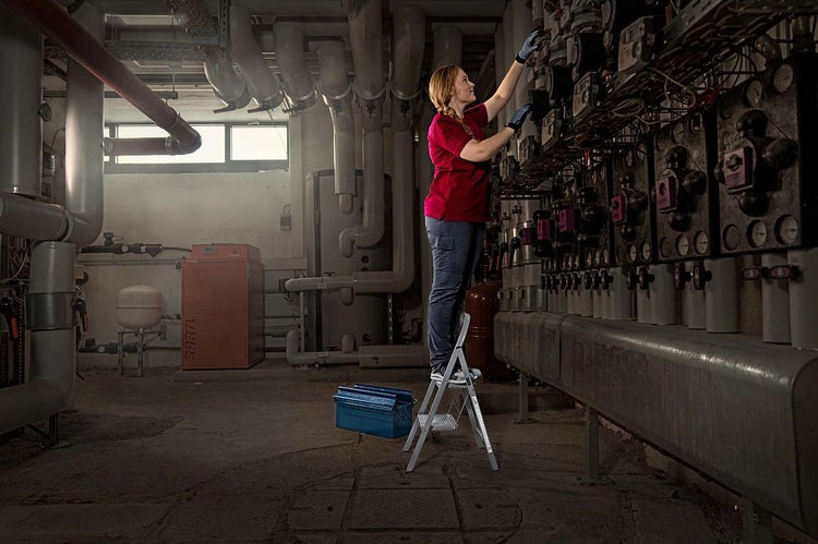 Eine Frau steht auf einer Trittleiter in einem Heizungsraum und arbeitet an der Heizungsanlage.