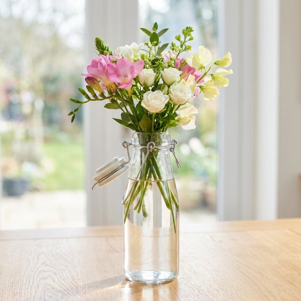 Ein Blumenstrauß aus Rosen, Freesien und Duftwicken in einer gläsernen Bügelflaschenvase auf einem Holztisch vor einem Fenster.