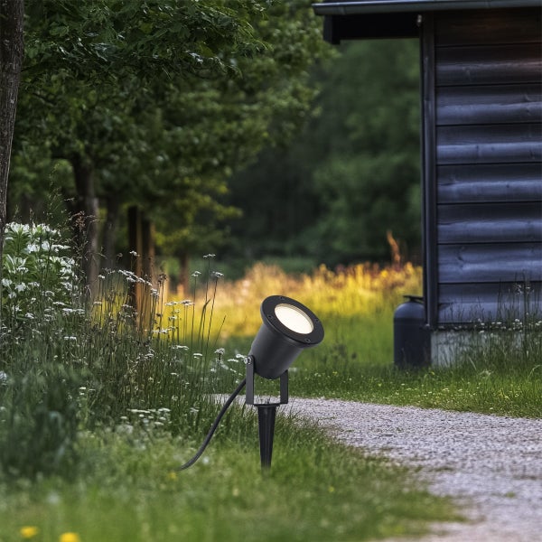 Schwarzer LED-Gartenstrahler mit Erdspieß beleuchtet einen Kiesweg im Garten neben einem Gartenhaus aus Holz.