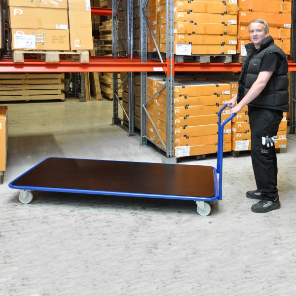 Großer Plattformwagen mit blauem Stahlrahmen und dunkler Holzfläche, geführt von einem Mitarbeiter in einem organisierten Lager mit Hochregalen.
