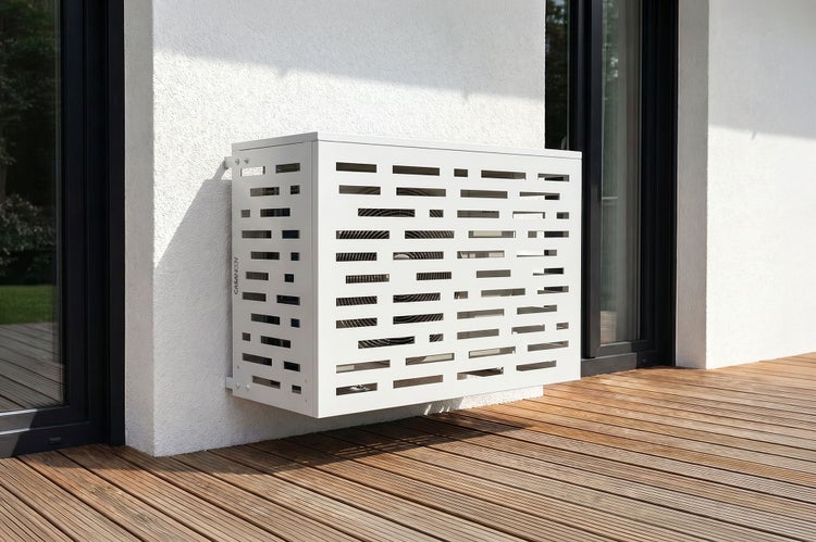 Weiße Klimaanlagen-Abdeckung aus Metall mit dekorativen Schlitzen, montiert an einer Hauswand über einer Holzterrasse.