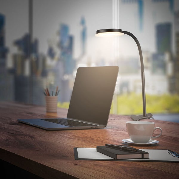 Moderne LED-Klemmleuchte auf einem Holzschreibtisch neben einem Laptop und einer Kaffeetasse in einem hellen Büro.