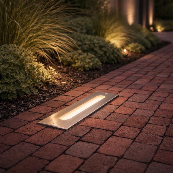 Rechteckige LED-Bodenleuchte aus Edelstahl, installiert in einem gepflasterten Gartenweg neben Pflanzenbeeten.