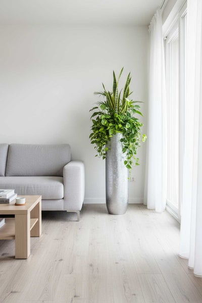 Helles, organisiertes Wohnzimmer mit hohem silbernem Pflanzkübel, grünen Zimmerpflanzen, grauem Sofa und Couchtisch aus hellem Holz.