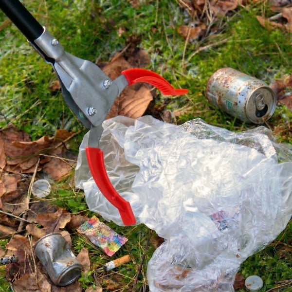 Müllgreifer aus Metall beim Aufsammeln einer Plastiktüte von einer Wiese mit Laub und Abfall.