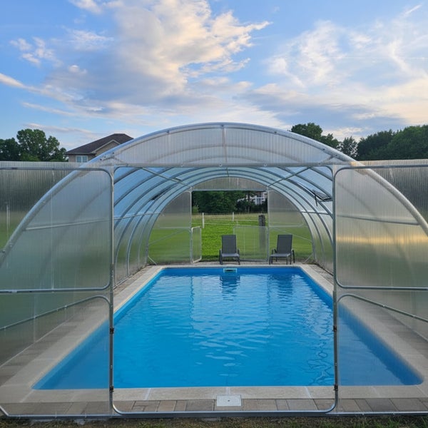 Rechteckiger Pool im Garten mit einer halbkreisförmigen, transparenten Poolüberdachung aus Kunststoff und Metallrahmen, zwei Liegestühle am Ende.