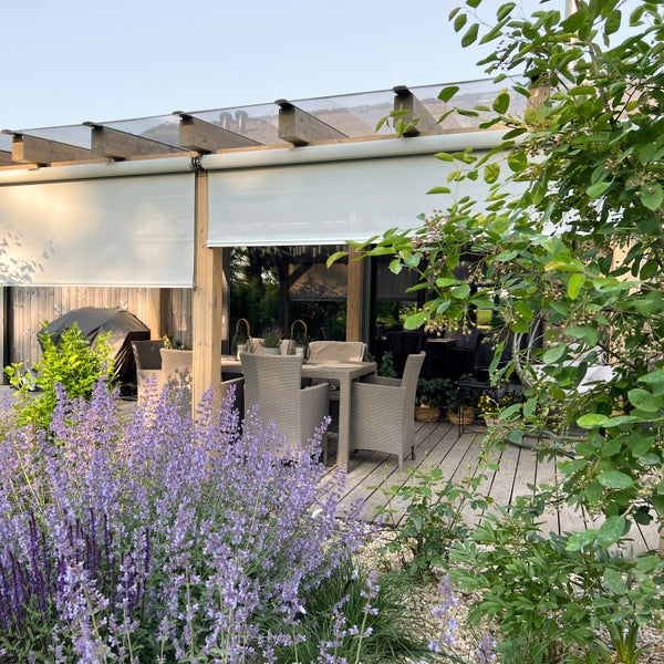 Moderne Holzterrasse mit Esstisch, grauen Sesseln und einer Pergola mit weißen Rollos unter einem Glasdach, umgeben von lila Blumen im Garten.