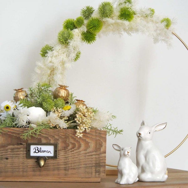 Dekorative Blumengesteck in Holzkiste mit Hasenfiguren und goldenem Ring
