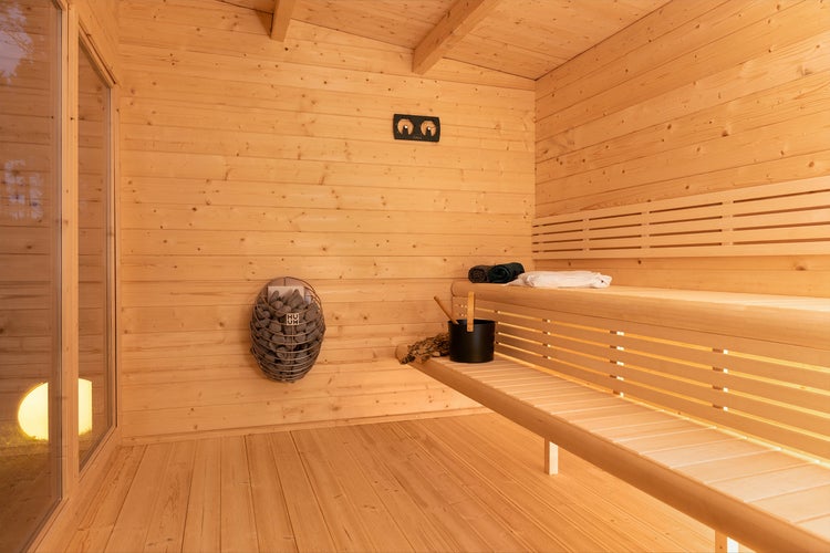 Innenansicht einer hellen Holzsauna mit mehrstufigen Bänken, wandmontiertem Saunaofen mit Steinen, Saunaeimer und indirekter Beleuchtung.
