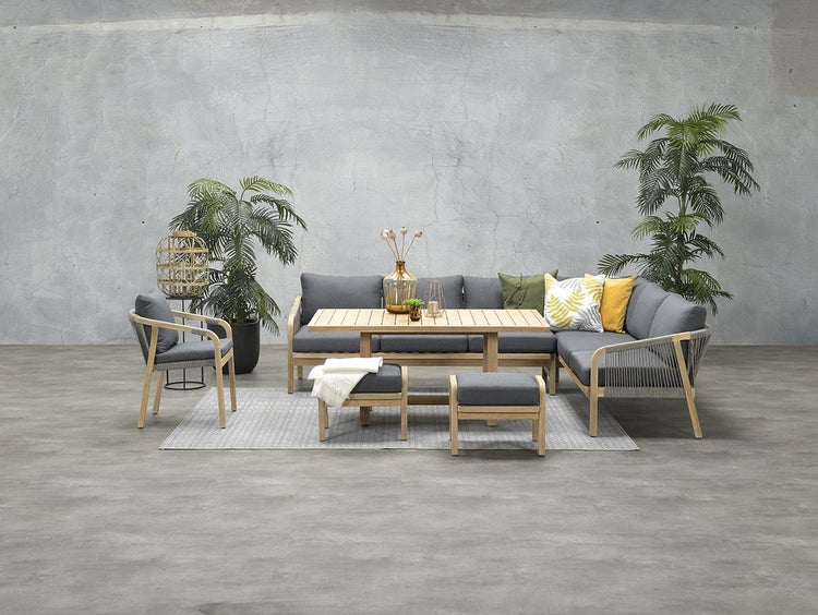 Modernes Garten-Lounge-Set aus Holz mit Ecksofa, Sessel, Hockern und Tisch, graue Polster, Seilbespannung, Pflanzen und Kissen in einem hellen Raum.