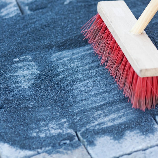 Besen mit roten Borsten kehrt dunklen Fugensand in die Fugen von Pflastersteinen ein.