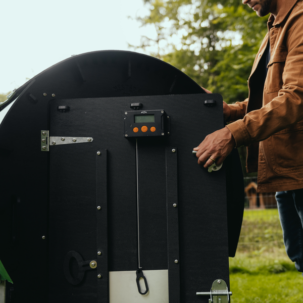 Person bedient einen automatischen Hühnerklappenöffner mit digitalem Display an einem dunklen Hühnerstall aus recyceltem Kunststoff im Außenbereich.