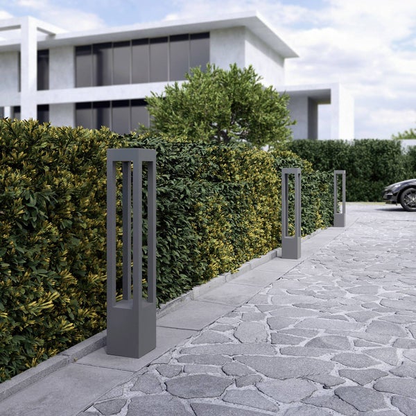 Drei moderne anthrazitfarbene Pollerleuchten an einer gepflasterten Einfahrt neben einer grünen Hecke vor einem hellen modernen Gebäude.