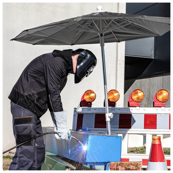 Ein Schweißer mit Schutzkleidung und Schweißhelm arbeitet unter einem großen schwarzen Baustellenschirm im Freien.
