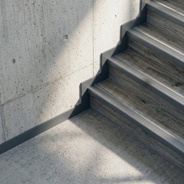 Graues Sockelleistenprofil an einer Treppe mit Holzoptik-Stufen und einer Sichtbetonwand in einem hellen Raum.