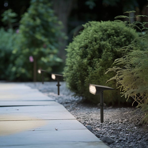 Schwarze LED-Gartenstrahler mit Erdspieß beleuchten einen grauen Steinweg und grüne Sträucher in einem hellen Garten.