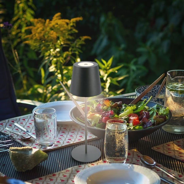 Akku-LED-Tischleuchte in Dunkelgrau auf einem gedeckten Gartentisch neben einer Salatschüssel im Freien.