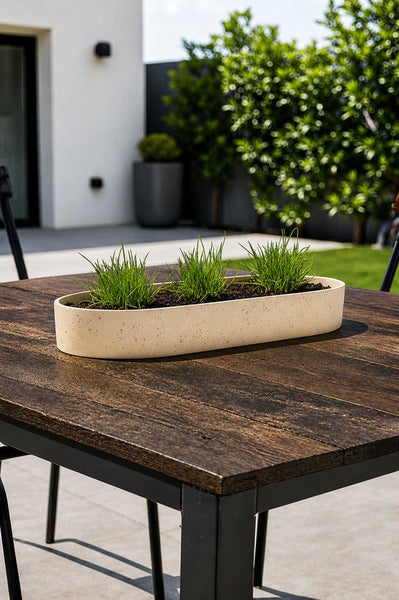 Ovales, gesprenkeltes Pflanzgefäß mit drei Pflanzen auf einem Holztisch im Außenbereich vor einer Hauswand und Hecke.