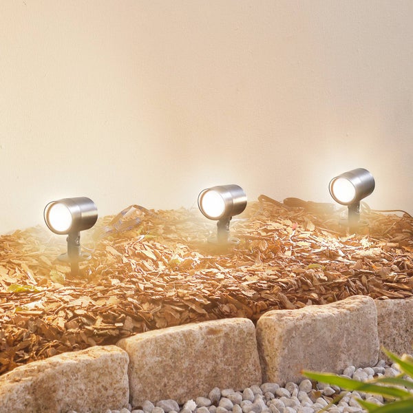 Drei eingeschaltete Gartenstrahler aus Metall in einem mit Rindenmulch gefüllten Beet mit Steineinfassung.