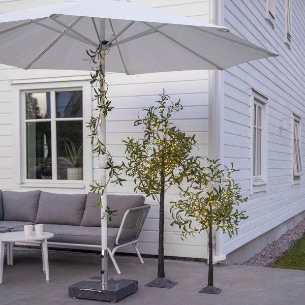 Außenbereich mit Sonnenschirm, Lounge-Möbeln und dekorativen Olivenbäumen mit Beleuchtung.