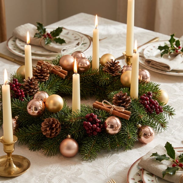 Festlicher Adventskranz mit Tannenreisig, Weihnachtskugeln, Tannenzapfen, Zimtstangen und vier brennenden Stabkerzen auf einem gedeckten Tisch.