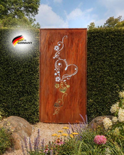 Rechteckiger Garten Sichtschutz aus Metall in Edelrost Optik mit ausgestanztem Herz und Blumen Motiv vor einer Hecke platziert Made in Germany