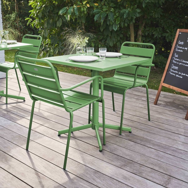 Grüner quadratischer Gartentisch und zwei Stühle aus Metall auf einer Holzterrasse, umgeben von Pflanzen.
