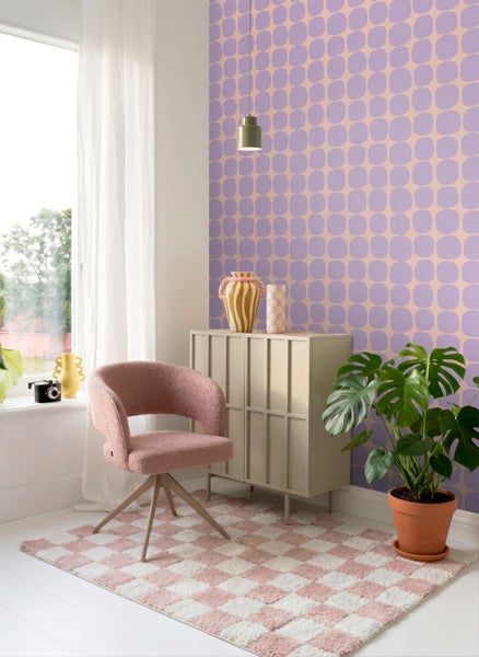 Hell gestalteter Raum mit gemusterter Tapete, rosa Sessel, beige Kommode, kariertem Teppich und einer Monstera Pflanze.