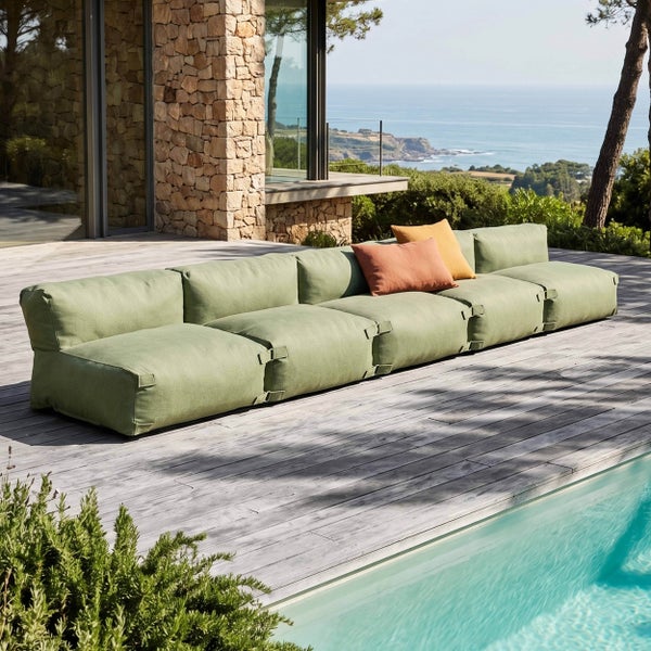 Modulares Lounge-Sofa in Salbeigrün auf einer hellen Holzterrasse am Pool mit weitläufigem Meerblick und Natursteinwand.