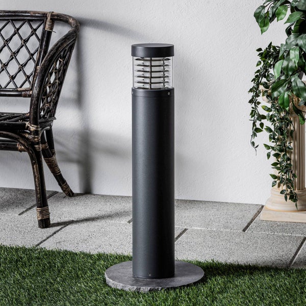 Moderne zylindrische Außenleuchte in Anthrazit auf einer Terrasse mit Steinplatten, Kunstrasen, einem Rattanstuhl und einer Pflanze.