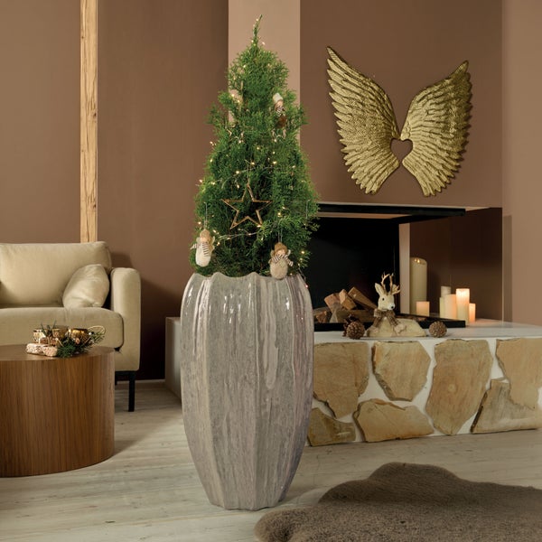 Weihnachtlich geschmückte Konifere in einem modernen, hohen Pflanzkübel in einer gemütlichen Wohnzimmerkulisse mit Kamin und Weihnachtsdekoration.