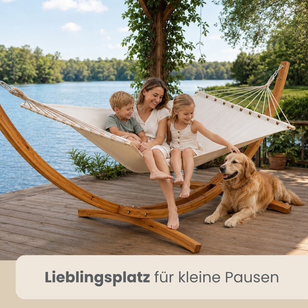 Große Hängematte mit Holzgestell auf einer Holzterrasse am See mit einer Frau, zwei Kindern und einem Hund in heller Umgebung.
