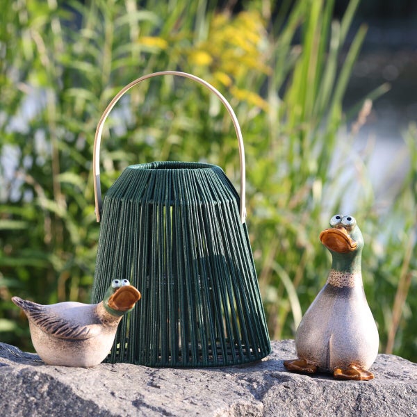 Gartenlaterne aus Kordelgeflecht mit Henkel und zwei dekorative Entenfiguren aus Keramik auf einem Stein im Garten