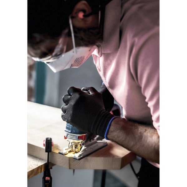 Eine Person mit Schutzbrille und Mundschutz sägt mit einer Stichsäge ein Holzbrett.