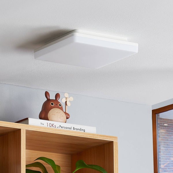 Moderne, flache quadratische LED-Deckenleuchte in einem hellen Raum über einem Holzregal.