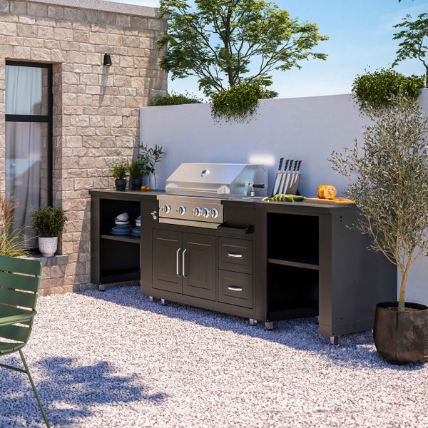 Moderne Outdoorküche mit integriertem Edelstahl-Gasgrill, Schränken und Ablageflächen auf einer Kiesterrasse vor einer Steinmauer.