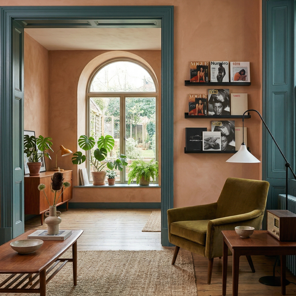 Wohnzimmer mit pfirsichfarbenen Wänden, blaugrünem Türrahmen, grünem Samtsessel, Holzmöbeln, Zimmerpflanzen und einem großen Rundbogenfenster.